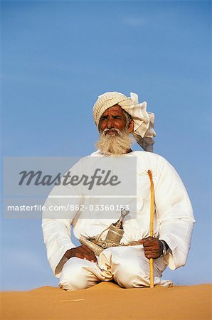 Un homme de bédouins s'agenouille au sommet d'une dune de sable dans le désert. Il porte la traditionnelle omanaise blanc manteau ou plat dash, un turban, un cérémonial courbé dague (khanjar) et détient une courte stickOman chameau 2 JWLJohn Warburton-Lee
