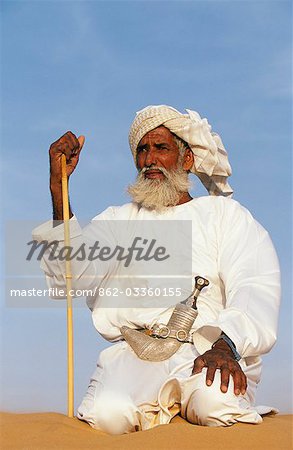 A Bedouin man kneels on top of a sand dune in the desert . He wears the traditional Omani long white cloak or dish dash,a turban,a ceremonial curved dagger (khanjar) and holds a short camel stickOman 7John Warburton-Lee