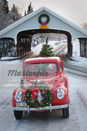 Man driving a vintage 1941 Ford pickup through a covered bridge with a Christmas wreath on the grill and a tree in the back during Winter in Southcentral, Alaska