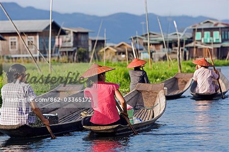 Myanmar,Burma,Lake Inle. A group of Intha women paddling their flat-bottomed wooden boats to market with typical Intha houses on stilts in the background.