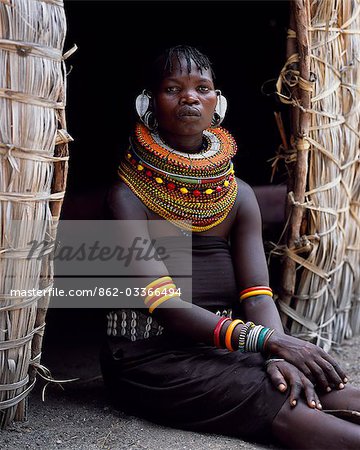 A Turkana woman,typically wearing many layers of bead necklaces and a series of hooped earrings with an pair of leaf-shaped earrrings at the front,sits in the entrance to her hut.