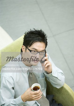 Businessman sitting in armchair, using cell phone and holding cup of coffee