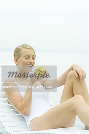 Teenage girl wearing bathing suit, sitting in lounge chair, eating apple