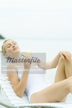 Teenage girl wearing bathing suit, sitting in lounge chair, holding slice of pineapple
