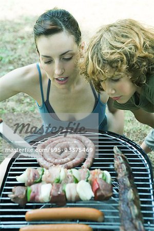 Young woman and boy bending over barbecue, looking at grilling meats