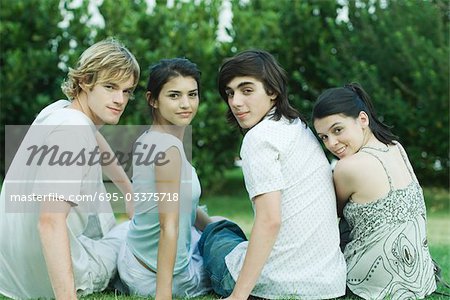 Four young friends sitting on grass, side by side, looking over shoulders at camera