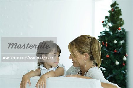 Mädchen und Mutter auf Sofa lächeln einander, Weihnachtsbaum im Hintergrund