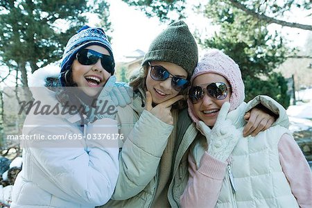 Three teen girls wearing winter clothes and sunglasses, holding chins, laughing