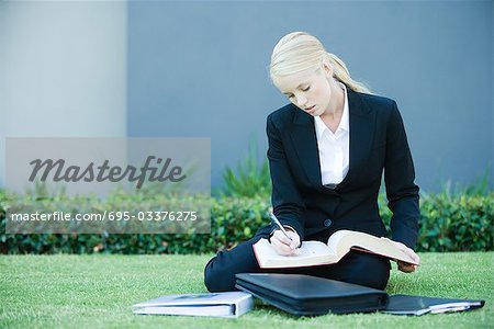 Jeune femme assise sur le sol en plein air, livre de lecture, tenant le stylo