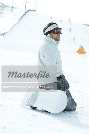 Young snowboarder kneeling on ski slope, looking away, side view