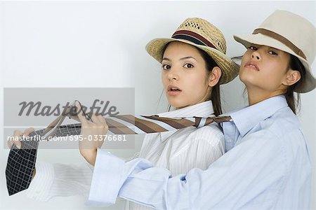 Two young female friends dressed in hats and ties, one behind the other, looking at camera