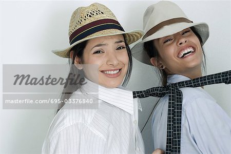 Two young female friends dressed in button down shirts, ties and hats, one pulling the other's tie, laughing, portrait