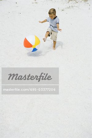 Young boy kicking beach ball at the beach, arms out