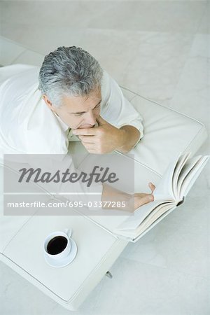 Homme mûr, allongé sur la chaise longue avec la main sous le menton, livre de lecture, vue d'angle élevé