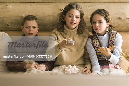 Three young siblings watching TV, teen girl changing channel while brother and sister complain