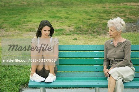 Teenage girl sitting apart from grandmother on bench, both frowning