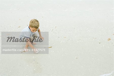 Little boy squatting, picking up ball on beach, full length