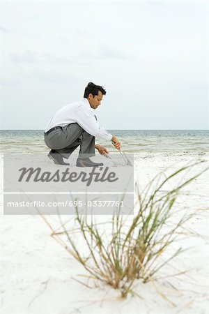 Homme accroupi sur la plage, balayant le sable en pelle, vue latérale