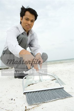 Homme accroupi sur la plage, balayant le sable en pelle, regardant la caméra, vue d'angle faible