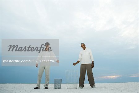 Two men at the beach holding their hands over garbage cans, one looking at camera
