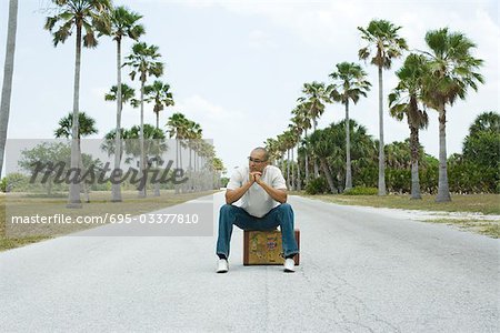 Man sitting on suitcase in the middle of street, looking away