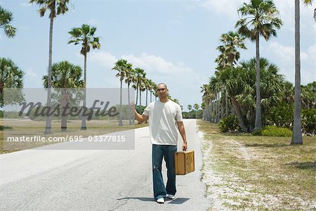 Man hitchhiking beside road, carrying suitcase