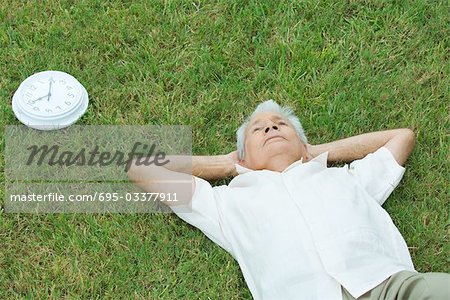 Senior homme couché sur l'herbe suivant pour l'horloge, les mains derrière la tête, grande angle vue