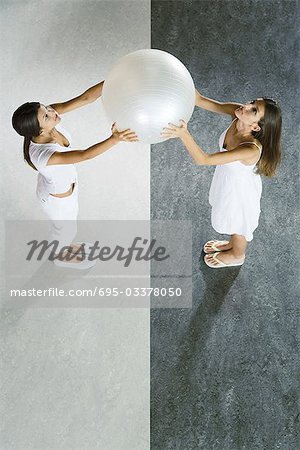 Teenage twin sisters standing face to face, holding up fitness ball together, viewed from directly above