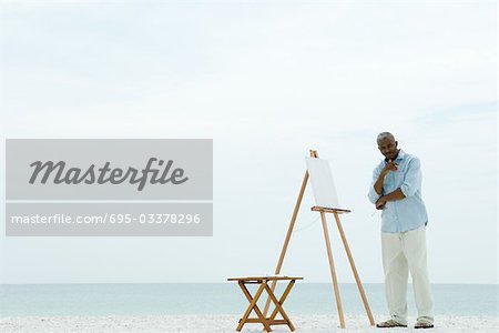 Senior homme debout à côté de la toile blanche à la plage, l'utilisation des pinceaux, souriant à la caméra
