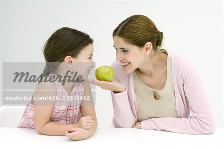 Mother and daughter sitting face to face, smiling at each other, woman holding apple