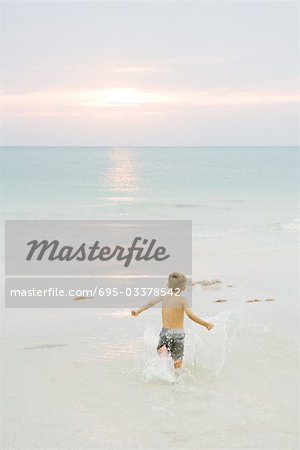 Little boy running at the beach, arms out, rear view