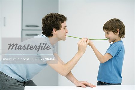 Father and son standing face to face, holding up green beans between their mouths, side view