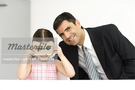 Father and daughter sitting side by side, girl looking through slice of bread with heart-shaped holes, both smiling at camera