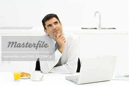 Homme d'affaires assis à table avec ordinateur portable et le petit déjeuner, la main sous le menton, en levant