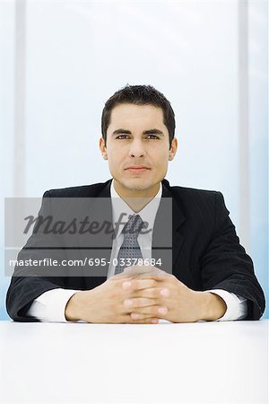 Businessman sitting at table with clasped hands, looking at camera