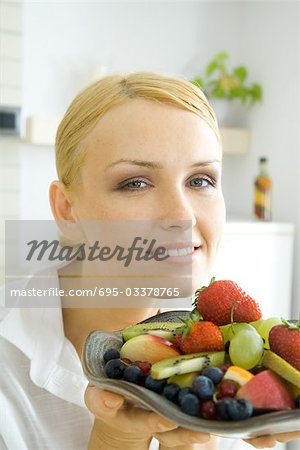 Woman holding assorted fruits on plate, smiling at camera