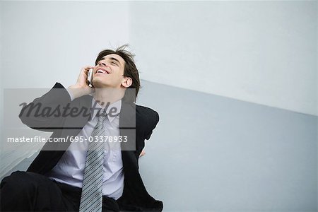 Junger Mann im Anzug am Boden sitzend mit Handy, Kopf nach hinten