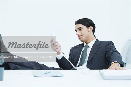 Young businessman sitting at desk with feet up, looking at landline phone, smiling