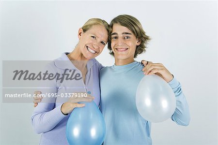 Mère et fils adolescent avec bras autour des épaules de l'autre, tenant des ballons, souriant à la caméra