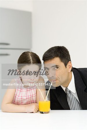 Father and daughter sharing glass of orange juice, drinking from straws, looking at camera