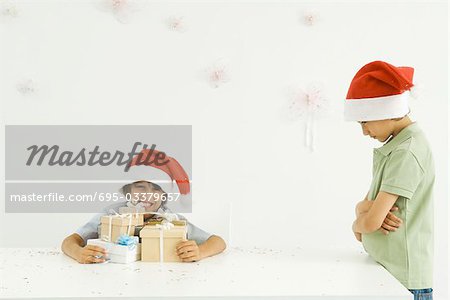 Boy holding stack of Christmas gifts, brother standing nearby, sulking, both wearing Santa hats