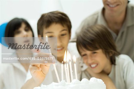 Boy placing burning candles on birthday cake, family watching