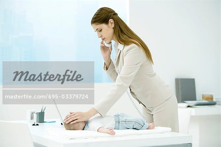 Young working mother in office, baby lying on desk, woman stroking baby's head and making phone call