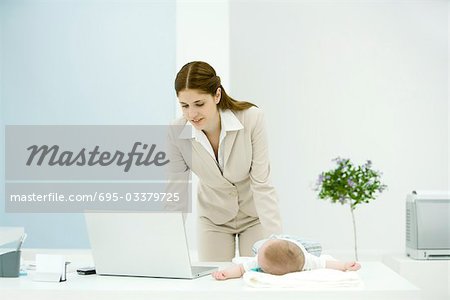 Young working mother using laptop, baby lying on desk