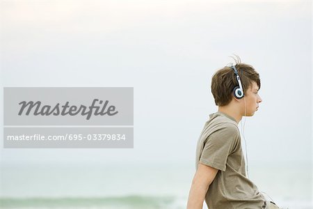 Jeune garçon assis sur la plage de l'écoute au casque, vue latérale