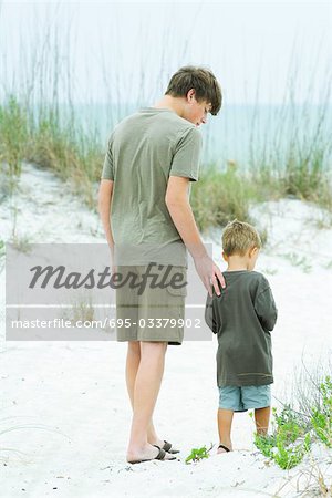 Teen boy walking with little brother at the beach, rear view