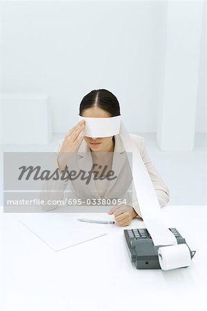 Woman sitting at desk, tape from an adding machine wrapped around her eyes, touching face
