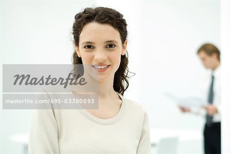 Young professional woman smiling at camera, colleague in background