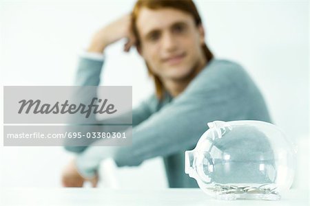 Transparent piggy bank sitting on table, young man in background holding head