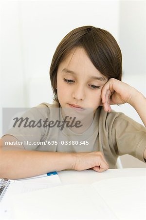 Boy sitting at table looking at homework, frowning